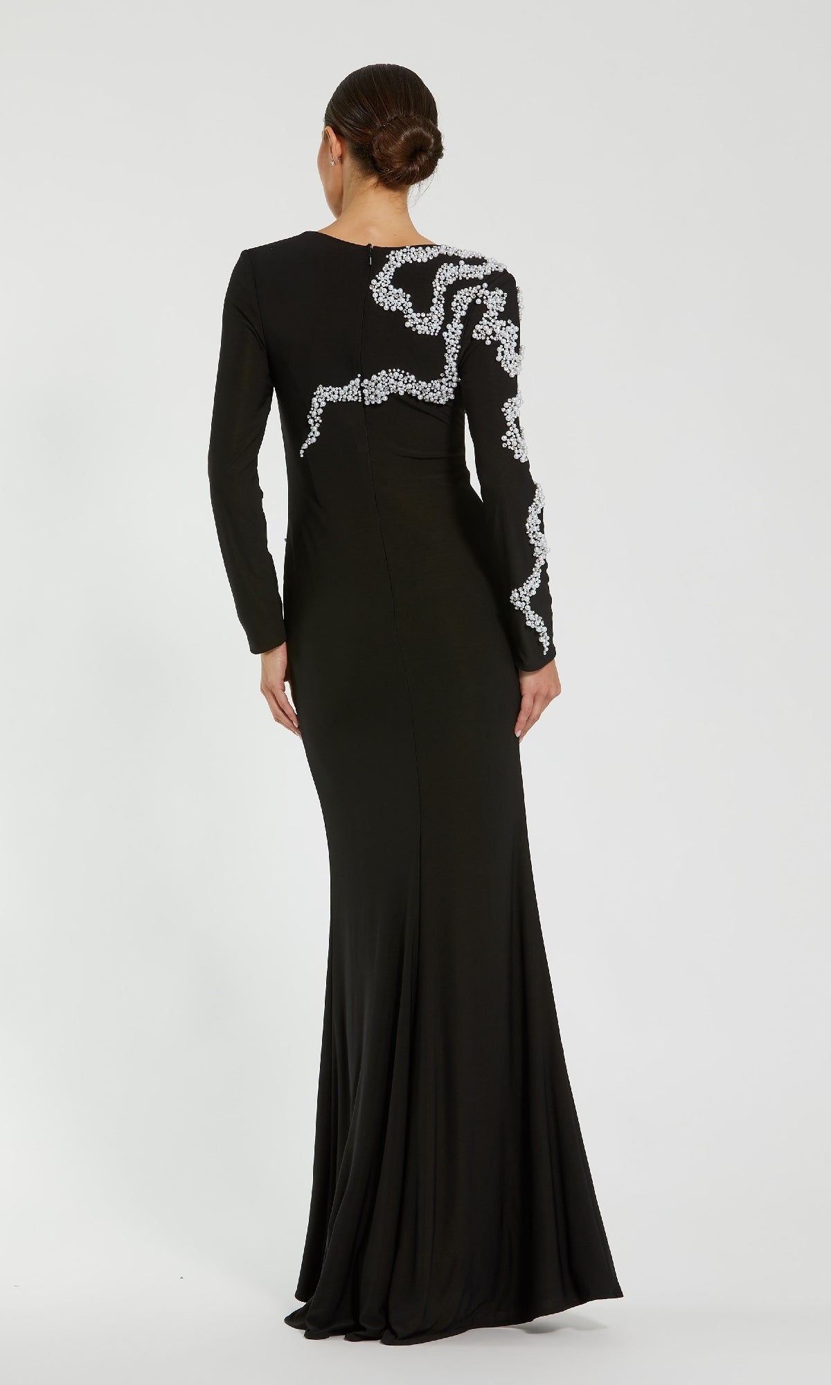 A woman faces away, wearing the Mac Duggal 44086 long formal dress—an elegant black jersey evening gown with long sleeves and white beaded details on the back and one sleeve. Her hair is styled in a low bun.